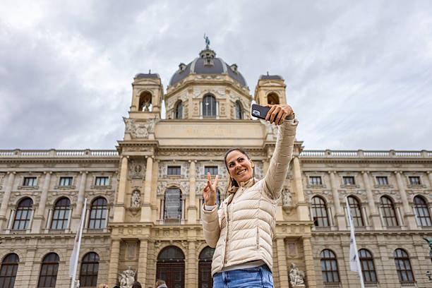 A tourist is taking a selfie in front of a beautiful historical building during a cloudy day. The architecture is stunning, with detailed facades and towering structures