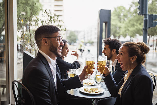 A cheerful group of business colleagues enjoy drinks and food at an outdoor cafe, celebrating and toasting in casual attire.
