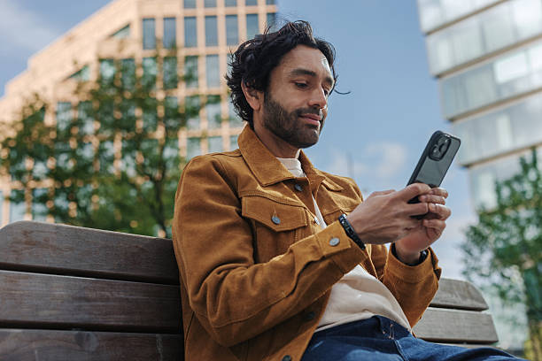 Young adult man sitting on a city bench, using a smartphone to browse social media and connect with friends in a sunny urban setting