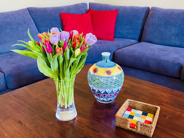 A glass vase with tulip flowers that are different colors orange, dark red (or purple) and pink on a wood brown coffee table. Also a colorful porcelain vase and wood holder for napkins that are colorful too. In the background is blue couch with 2 red pillows. Located in California during Summer Time 2025.
