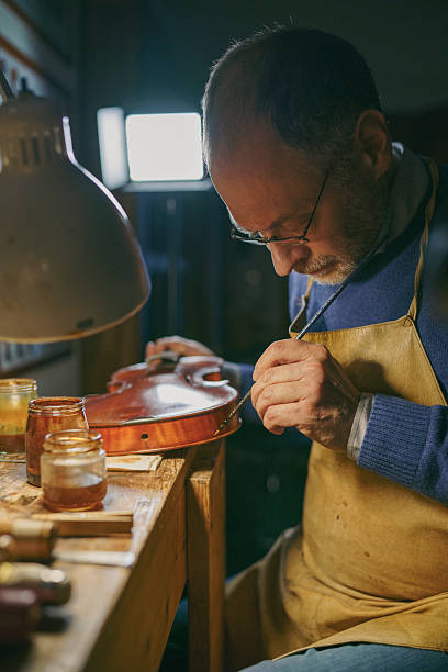 istock Skilled luthier carefully applying varnish to violin, surrounded by woodworking tools in traditional craftsman workshop, highlighting meticulous instrument-making process 2221064520