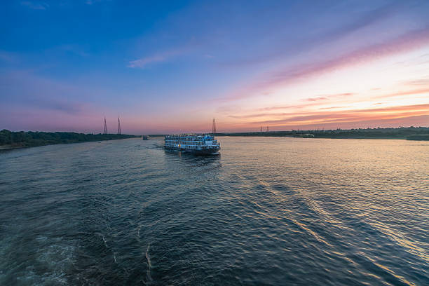 A large cruise ship glides along the tranquil Nile River near Esna Lock, Aswan, Egypt, at dusk. The sky above is painted with soft purple and pink hues, reflecting on the water.