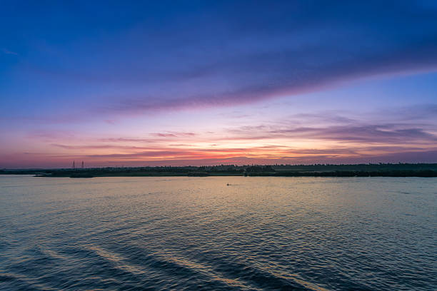 A tranquil wide shot of the Nile River at sunset near Esna Lock, Aswan, Egypt. The sky displays soft pink and purple hues, with silhouetted power towers and palm trees along the distant riverbanks.