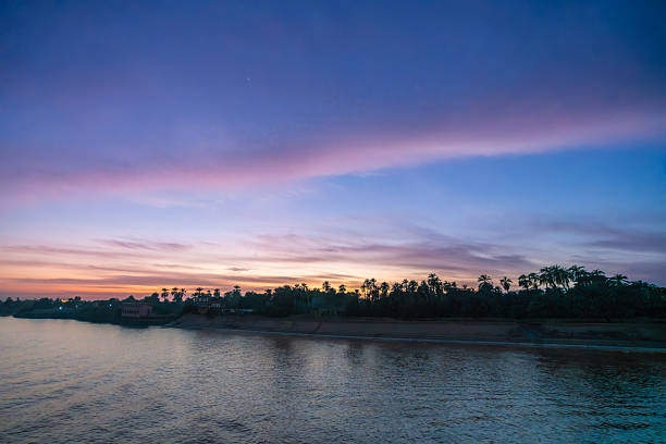 A captivating dusk scene on the Nile River in Aswan, Egypt, as vibrant pink and purple hues fill the sky. Lights from local houses illuminate the riverbank, with silhouettes of palm trees.