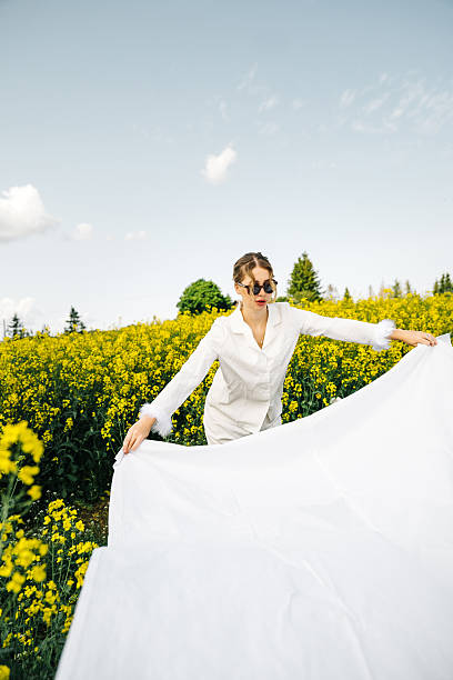 This evocative image depicts a young woman with curly red hair walking through a sunlit meadow during late afternoon. Her vintage-style white dress, with puffed sleeves and a tie at the neck, adds a timeless elegance to the scene. She appears absorbed in the moment, gently touching the wildflowers as she walks by. The warm, natural lighting and the open field background enhance the pastoral and peaceful ambiance, evoking a sense of freedom and connection to nature.