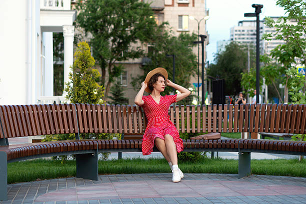 Woman adjusts straw hat while seated on park bench outdoors.