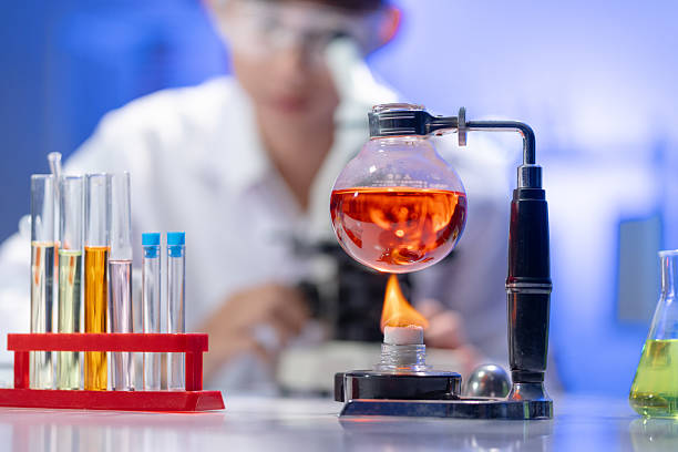 A flask of red liquid is heated over a flame on a lab bench filled with equipment. In the background, a blurred scientist works, creating a scene of active research and development.