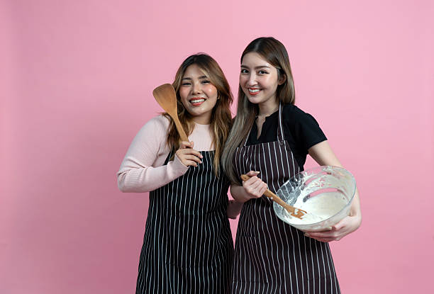 istock Two women are having fun baking together. They are wearing apron and smiling while mixing ingredient with a wooden spoon. Portrait on pink background with studio light. 2221109182