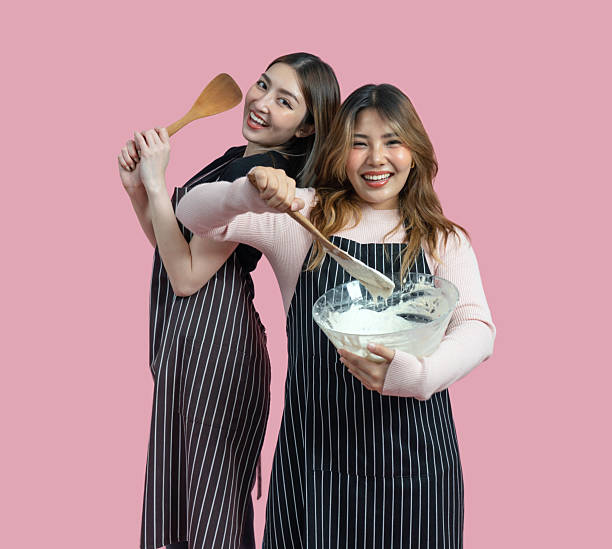 istock Two women are having fun baking together. They are wearing apron and smiling while mixing ingredients with a wooden spoon. Portrait on pink background with studio light. Isolated 2221109184