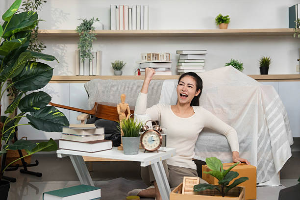 A delighted woman expressing joy as she unpacks boxes in her new living space, surrounded by plants and decor.