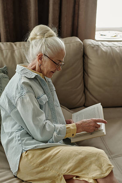 istock Elderly Woman Reading Book on Beige Sofa 2221113112