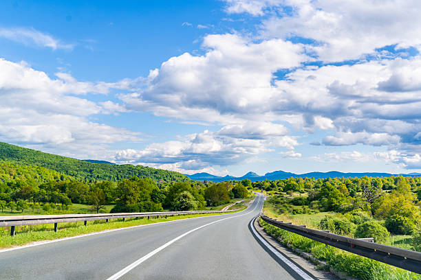 Smooth asphalt road bordered by guardrails and green hills, curving gently into the distance. The sky is bright blue with a few scattered clouds, enhancing the open, sunny atmosphere of this rural landscape in Croatia.