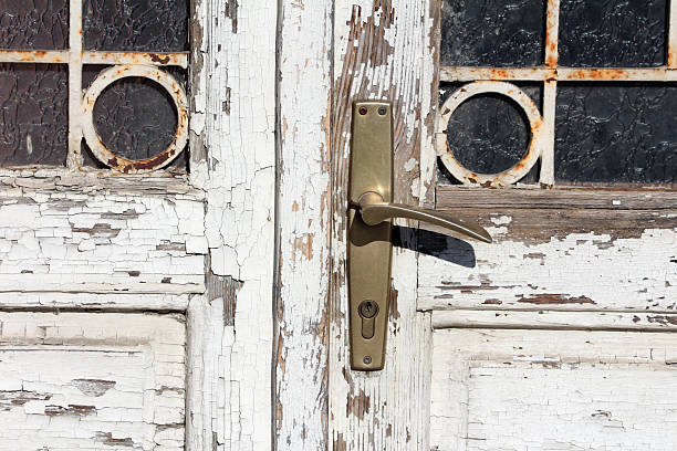 Close-up of an old white wooden door with peeling paint, brass handle, and stained glass panels with rusty circular motifs, showing age and texture in bright sunlight