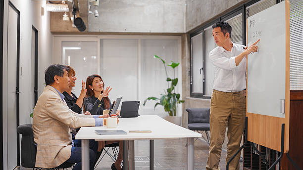 A deaf businessman is using a whiteboard and teaching sign language to his colleagues in a modern office. His colleagues are very eager and enthusiast to learn sign language. There is also a non-binary business person in the team.