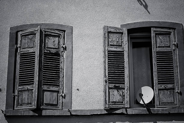 Two vintage wooden shutters hang wide open beside a window in an old village home. The weathered textures tell stories of time, complemented by a satellite dish reflecting a modern touch.