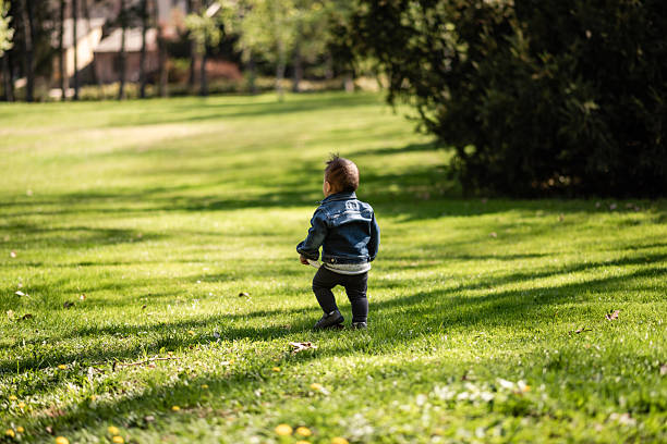 Young child walking away from camera on grassy lawn in park wearing denim jacket, exploring nature and enjoying outdoor playtime