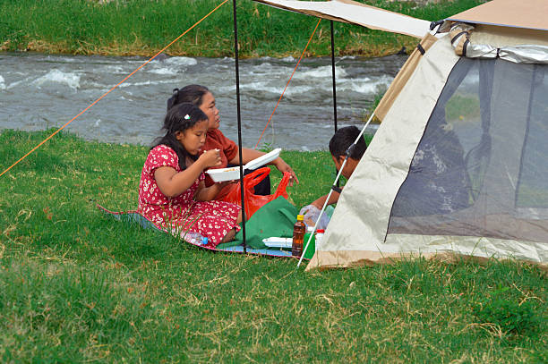 Two mothers and their kids sit comfortably near the tent, chatting and enjoying lunch in a relaxed and cheerful setting