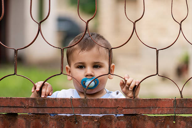 Pensive toddler with a pacifier gazing through a rusty metal fence in the backyard, reflecting curiosity and a sense of longing