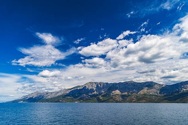Wide-angle panoramic view of the Croatian coastline as seen from a ferry crossing the Adriatic Sea. The composition captures a dramatic sky with scattered white clouds above the rugged mountains of Dalmatia, contrasted by the calm, deep blue water below. This high-resolution image is ideal for travel promotions, landscape themes, and backgrounds showcasing the natural beauty of coastal Croatia.