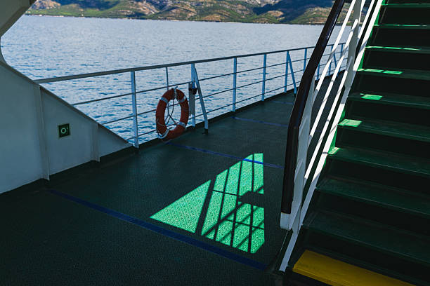 A creative composition showing a quiet corner of a Croatian ferry deck, where the sunlight casts geometric shadows through the ship’s railing onto the textured green floor. A red life ring hangs on the guard rail, while steep green stairs enter the frame on the right. The image captures a minimalist yet atmospheric moment during a summer voyage across the Adriatic, blending form, function, and natural light.