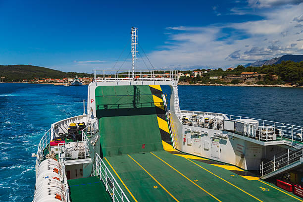 A high-angle view of an empty ferry deck as it sails away from the Croatian coast. The raised green ramp, yellow safety lines, and visible wake contrast beautifully against the deep blue of the Adriatic Sea. Ideal for illustrating summer marine transportation, ferry logistics, or coastal travel in Europe.
