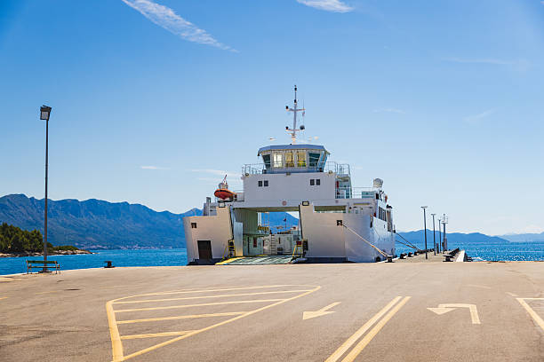 A large white ferry stands docked on the Croatian coast, ready for vehicle and passenger boarding. The open ramp and wide asphalt loading zone are set against a dramatic backdrop of blue sea, mountainous shoreline, and cloudless sky, creating a calm and professional maritime atmosphere ideal for depicting transport, travel, or infrastructure themes.