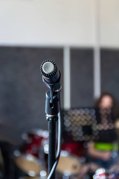 A microphone stands in focus at a music rehearsal in a studio. Musicians are playing instruments in the background, showcasing a creative and dynamic environment during the afternoon session.
