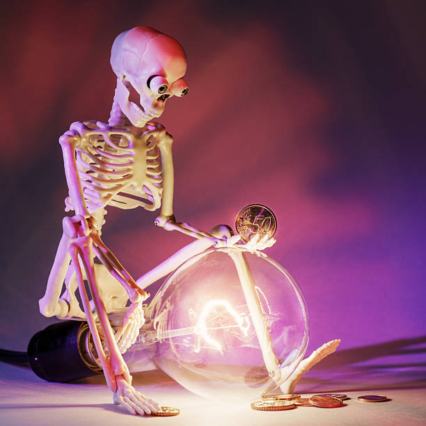 Surprised by the price of electricity, a toy model of a sad skeleton sitting on a glowing electric light bulb. Square photo.