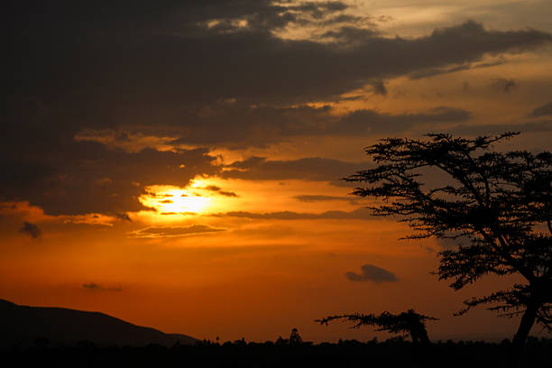 The sun breaks through dark clouds as it sets  against a fiery orange sky in Kenya. The shadows of fever trees and mountains border the lower frame.