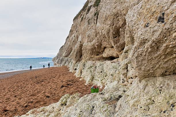 A beach with red sand meets a towering, layered cliff face under a cloudy sky in Butter Rock - Dorset - Great Britain