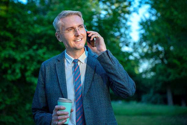 Smiling businessman talking on phone and holding coffee cup outdoors in park. stock photo