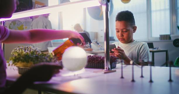 African American Elementary School Boy Sitting at Table with Girl, Growing Experimental Microgreens, Using Tweezer and Smartphone. Smart Young Students Studying Biology in Classroom. STEM Education.