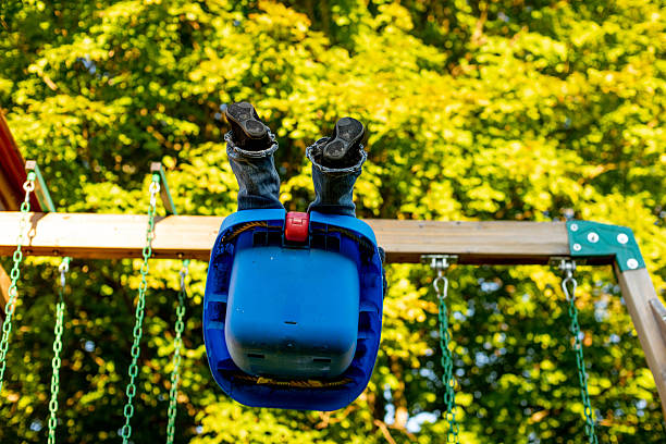 A young toddler enjoying playtime on a bright blue plastic swing set in a backyard surrounded by lush green trees and foliage. The image captures childhood joy, outdoor play, and family recreation activities.