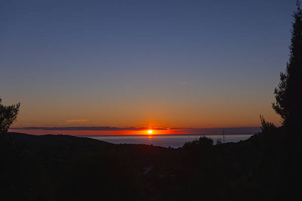 A calm and radiant sunset captured from an elevated viewpoint on Hvar Island, Croatia. The golden sun gently dips below the horizon, casting a vivid orange glow across the sea and the distant sky, framed by silhouettes of Mediterranean trees and low hills. This photograph reflects the tranquil essence of summer evenings in Dalmatia, offering ideal use for travel, nature, or inspirational editorial content.