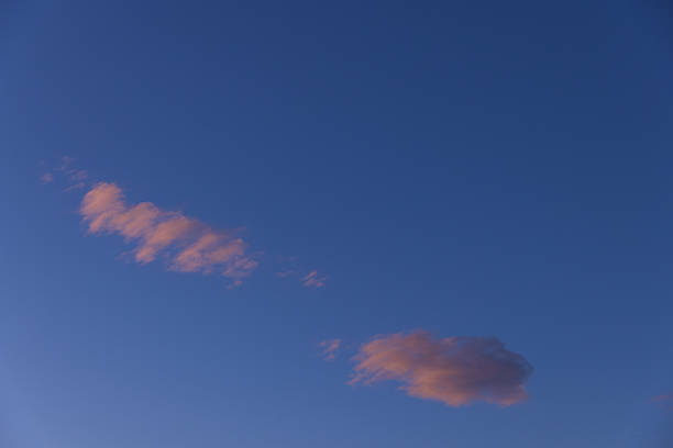 A minimalist evening sky photograph featuring two pink-hued clouds softly lit by the setting sun, drifting through a deep blue backdrop over Hvar Island, Croatia. The warm pastel tones contrast beautifully with the clear atmosphere, evoking feelings of calm, clarity, and lightness. Perfect for themes related to simplicity, serenity, dreaminess, and tranquil nature moments.