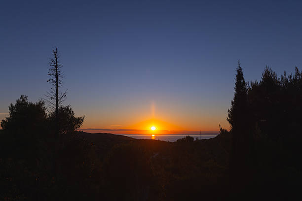 A breathtaking view of the sun setting over the Adriatic Sea, captured from an elevated point on Hvar Island, Croatia. The image features a vivid gradient sky, transitioning from deep blue to brilliant orange, as the glowing sun touches the horizon. Below, a coastal landscape with dark silhouettes of trees and hills is gently illuminated. A powerline tower subtly appears on the right, juxtaposing natural beauty with minimal human presence, enhancing the contrast between nature and infrastructure.