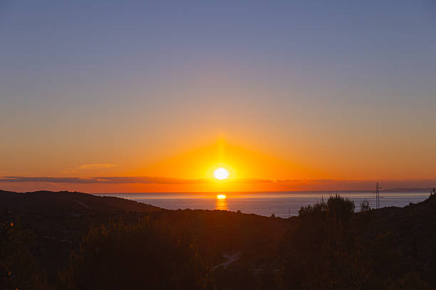 A breathtaking view of the sun setting over the Adriatic Sea, captured from an elevated point on Hvar Island, Croatia. The image features a vivid gradient sky, transitioning from deep blue to brilliant orange, as the glowing sun touches the horizon. Below, a coastal landscape with dark silhouettes of trees and hills is gently illuminated. A powerline tower subtly appears on the right, juxtaposing natural beauty with minimal human presence, enhancing the contrast between nature and infrastructure.