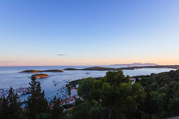 A breathtaking elevated view of Hvar’s coastline at golden hour, overlooking the vibrant marina filled with yachts, sailboats, and smaller vessels. The scene extends out to the calm Adriatic Sea with Pakleni Islands softly glowing under the warm sunset light. The foreground is partially framed by trees and red-roofed buildings of Hvar Town, adding depth and character to this tranquil coastal composition. A perfect depiction of Croatia’s maritime charm and summer tourism allure.