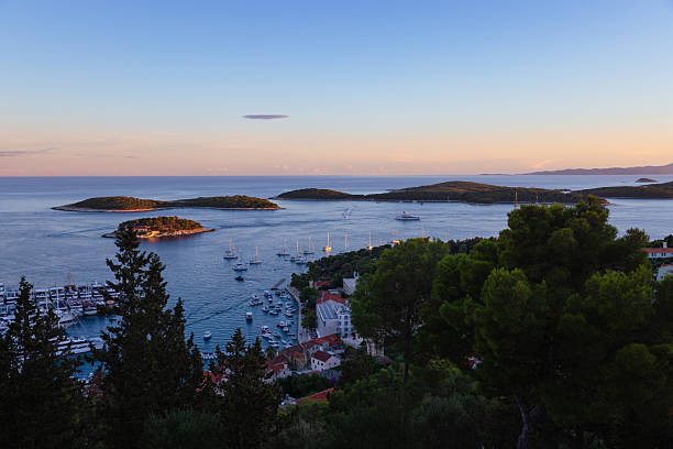 A breathtaking elevated view of Hvar’s coastline at golden hour, overlooking the vibrant marina filled with yachts, sailboats, and smaller vessels. The scene extends out to the calm Adriatic Sea with Pakleni Islands softly glowing under the warm sunset light. The foreground is partially framed by trees and red-roofed buildings of Hvar Town, adding depth and character to this tranquil coastal composition. A perfect depiction of Croatia’s maritime charm and summer tourism allure.