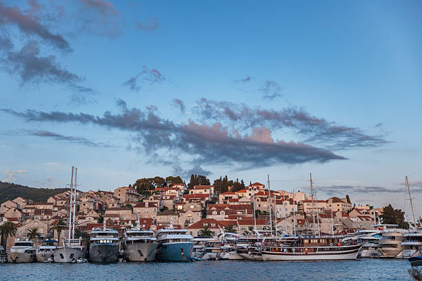 An atmospheric sunset image of Hvar’s waterfront, showcasing a picturesque blend of moored yachts, traditional Croatian boats, and stacked stone houses with red rooftops. The harbor is lined with elegant vessels, from sleek modern cruisers to classic wooden sailboats, all set against the backdrop of charming old town architecture nestled on a hillside. Wisps of dark evening clouds stretch across the pastel sky, enhancing the tranquil yet luxurious mood of this Adriatic gem.