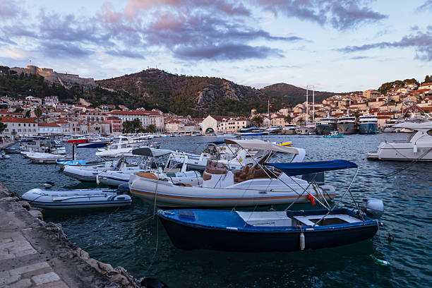 An atmospheric evening view of Hvar’s lively waterfront with small boats and speedboats anchored along the marina. In the background, the historic Fortica (Spanish Fortress) dominates the hilltop, while the charming old town of Hvar with its stone houses and terracotta roofs descends toward the sea. Tourists walk along the promenade, adding life to the picturesque Adriatic island scene during the summer golden hour.