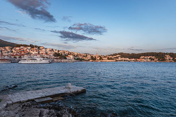 A picturesque view of Hvar Island’s vibrant harbor as the evening light bathes the historic Croatian town. The image captures elegant yachts anchored along the bustling waterfront, traditional stone houses with terracotta roofs rising in the background, and soft clouds drifting across a pastel blue sky. The calm Adriatic Sea laps against the stone jetty in the foreground, emphasizing the tranquil Mediterranean atmosphere of this iconic travel destination.