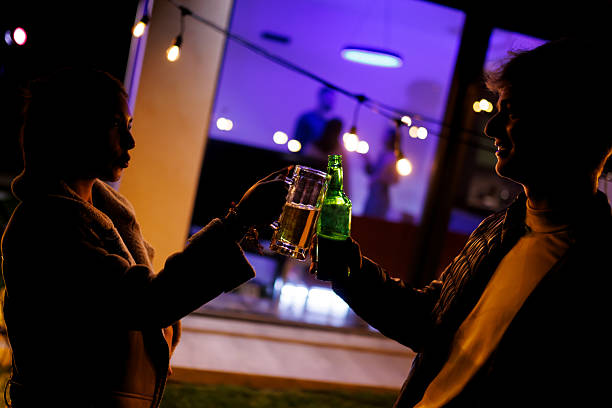 Silhouettes of two individuals toasting with drinks outdoors during a nighttime gathering, illuminated by festive string lights. Inside, a cozy ambiance with blurred party attendees