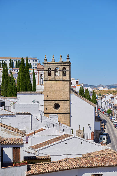 A scenic view of a rural Andalusian town, featuring whitewashed roofs and a historic bell tower. The clear blue sky enhances the tranquil charm of the Mediterranean architecture.