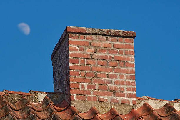 A brick chimney rises from a clay-tiled roof under a clear blue Danish sky, with the moon softly visible in the background. This scene showcases rustic architecture and serene evening light.