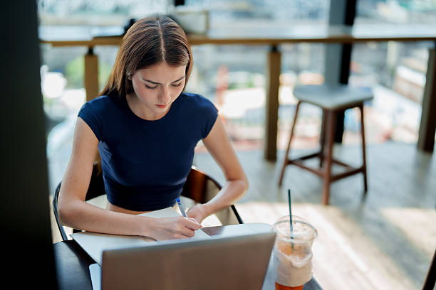 Focused young woman is working diligently her studies modern cafe, using laptop and taking notes notebook while enjoying refreshing iced drink. This scene captures essence of productivity