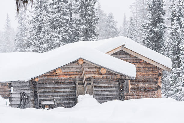 Two rustic wooden cabins are covered in thick snow while heavy snowfall creates a tranquil winter landscape in a forested area. The scene evokes warmth amid the cold.