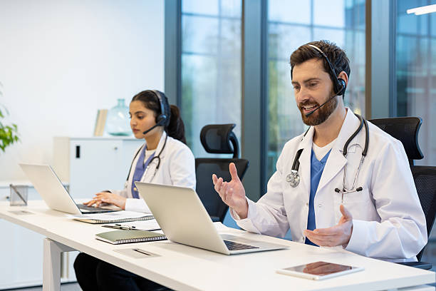 istock Two doctors working remotely consulting patients remotely, man and woman with headset using laptop for video call, working inside clinic, patient support and assistance service 2221277506