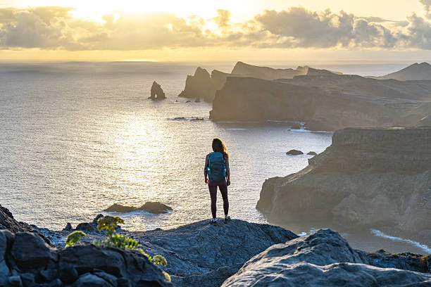 istock Female backpacker tourist enjoys panoramic view from view point on a steep cliff over the seascape and along the rugged foothills of Madeira coast at sunrise. Ponta do Bode, Madeira Island, Portugal, Europe. 2221303096