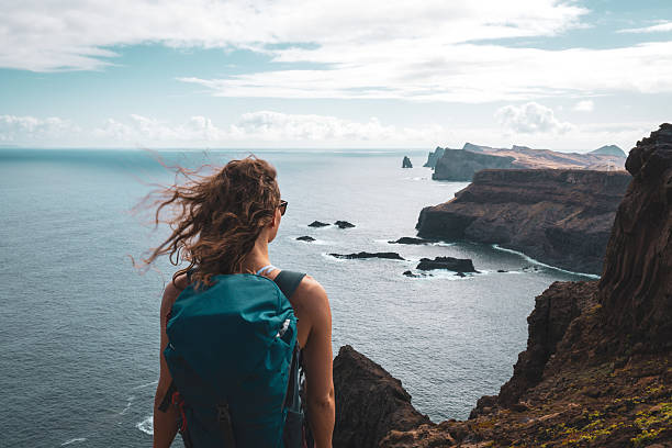 Description: Backpacker woman enjoys panoramic view from a steep cliff overlooking the sea and the rugged foothills of Madeira's coast in the morning. Ponta do Bode, Madeira Island, Portugal, Europe.